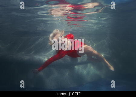 Blonde Frau unter Wasser im Pool, trägt ein rotes Kleid. Stockfoto