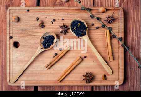 Schwarzer Tee mit Kräutern in Holzlöffel auf einem Holzbrett mit Zimt, Sternanis, Haselnuss und Pfeffer dekoriert. Oben in der Ansicht. Stockfoto