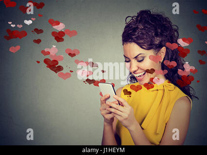 Schöne glückliche Frau senden Liebe SMS auf Handy mit roten Herzen fliegen weg von Bildschirm auf grauem Hintergrund isoliert. Positiven menschlichen e Stockfoto