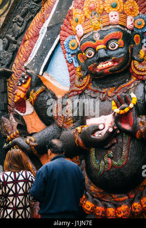 Ein Mann und eine Frau stellen Angebote am Fuße der Darstellung dieses Kala Bhairav in Kathmandu Durbar Square. Stockfoto