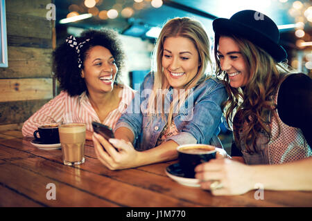 Junge Frau, die etwas auf ihrem Mobiltelefon mit zwei attraktiven lachende Mädchen Freunden zu teilen, wie sie sitzen zusammen in einem Café Kaffeetrinken Stockfoto