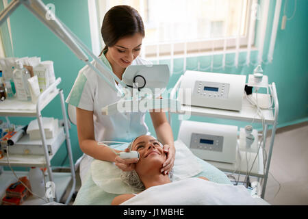 Junge Kosmetikerin unterziehen Kavitationsbehandlung auf Gesicht weiblich, Frau liegt und sanft lächelnd Stockfoto