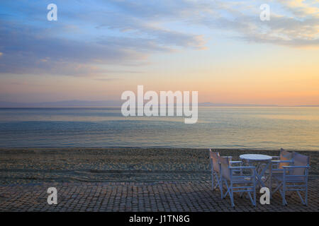 Tisch und Stühle in der Bar am frühen Morgen bei Sonnenaufgang des Tages mit einem ruhigen blauen Meer mit Blick auf den Himmel und die Berge des anderen Ufers, Kassandra, Gre Stockfoto