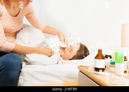 Mutter auf dem Bett sitzt und Fallenlassen Nasentropfen oder Spray zu ihrem kranken Kind Sohn Spritzen Stockfoto