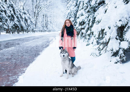 Siberian Husky sitzen in der Nähe von junge Frau, verschneiten Wald im Hintergrund. Niedliche Mädchen mit bezaubernden Hund Stockfoto