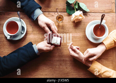 Weibliche und männliche Hände mit Ehering Draufsicht, Holztisch, Rose und Tassen auf Hintergrund. Paar romantisches Date. Heiratsantrag Stockfoto