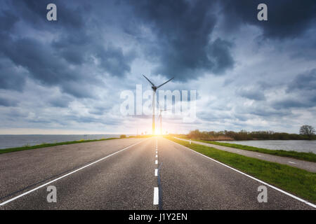 Schöne asphaltierte Straße mit Windkraftanlagen zur Stromerzeugung bei Sonnenuntergang. Windmühlen zur Stromerzeugung. Landschaft mit Strasse, grünen Rasen Stockfoto