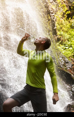 Wanderer-Mann aus ein natürliches frisches sauberes Wasser trinken. Junge Touristen Trinkwasser aus einer Flasche Outdoor-Wasserfall im Hintergrund. Trinkwasser, hydr Stockfoto