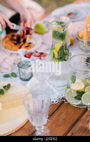 Picknick, veganes Essen, Ferienkonzept - lecker volle Krüge Limonade, in Scheiben geschnittenen Zitronen, Limetten, Minze, Glaswaren, Apfelkuchen, Kirschen und Eicheln auf festliche Holztisch mit weißen Tuch geschnitten. Stockfoto