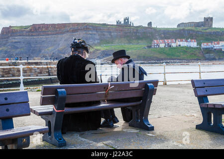 Ein Mann und eine Frau, gekleidet als Goten, machen Sie eine Pause, saß auf einer Bank am Pier bei Whitby, North Yorkshire, England, UK mit dem Kloster im Hintergrund Stockfoto