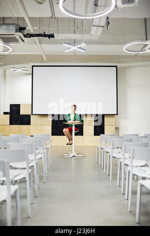 Junge Frau sitzt an Tisch im Hörsaal mit Whiteboard hinter Stockfoto