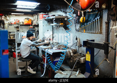 Seite Ansicht Porträt des modernen tätowierte Menschen Kontrolle defekten Motorteile am Tisch in der Motorradwerkstatt Stockfoto