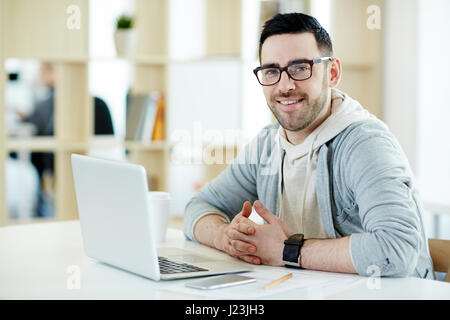 Porträt der moderne Mann mittleren Alters, lächelnd und Blick in die Kamera während der Arbeit mit Laptop im sonnendurchfluteten Büro Stockfoto