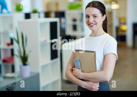 Porträt des jungen schöne Brünette Frau lächelt glücklich beim Betrachten der Kamera stehend im modernen Büro Stockfoto