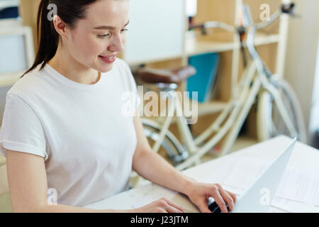 Porträt der hübsche junge Frau, gekleidet in Freizeitkleidung arbeiten mit Laptop und lächelnd in kreativen Büroräume Stockfoto
