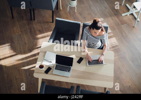 Draufsicht der jungen Frau am Schreibtisch sitzen und auf ihrem Laptop zu arbeiten, während das Telefon in der Nähe ist, und es auch einen anderen Laptop und Handy von ihrem Partner gibt. Stockfoto