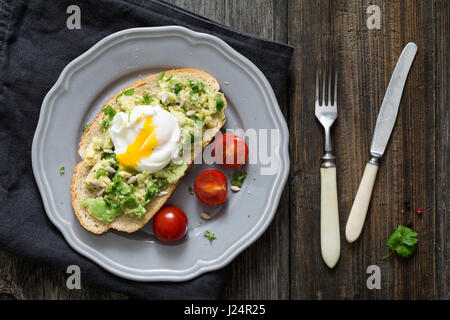 Gesundes Frühstück Toast: pochiertes Ei, pürierte Avocado mit Koriander und Limettensaft, Sonnenblumenkerne und Cherry-Tomaten auf einem Teller auf Holztisch Stockfoto