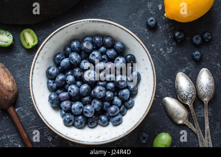 Heidelbeeren und Vintage Löffel: Draufsicht von frischen Heidelbeeren in weiße Keramikschale mit Baby Kiwi Frucht, Zitrone und Vintage Teelöffel auf dunklen Stein Stockfoto