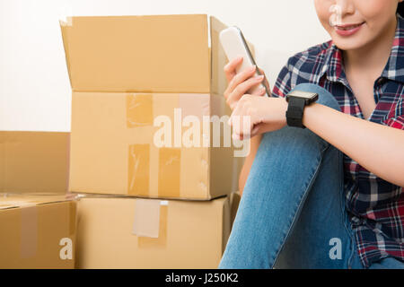 Frau mit Smartphone verbinden Smartwatch herumsitzen Kartons nach dem Umzug. Gemischte Rassen asiatische chinesische Modell. Stockfoto