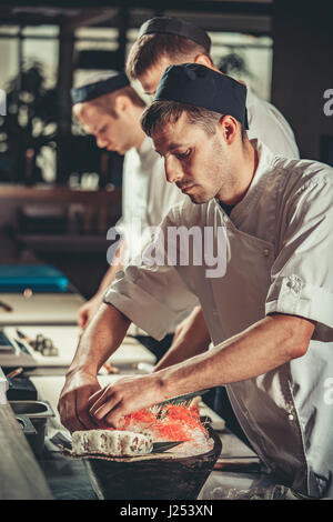 Drei junge Köche in weißer Uniform gekleidet dekorieren fertig Gericht im Restaurant. Sie arbeiten an Maki-Rollen. Traditionelles japanisches Sushi Zubereitung eingestellt Stockfoto