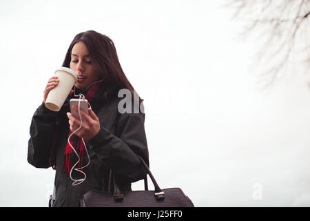 Glückliche Frau hören von Musik auf dem Handy beim Kaffeetrinken gegen Himmel Stockfoto