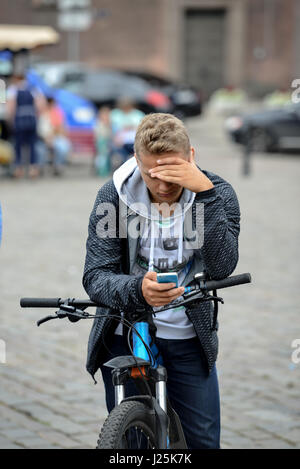 RIGA, Lettland - 27. Juli 2016: Unbekannter junger Mann auf einem Fahrrad auf der Straße angehalten und schaut auf das Telefon. Stockfoto