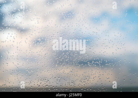 Regentropfen auf ein Fensterglas gegen blauen Himmel mit Wolken. Stockfoto