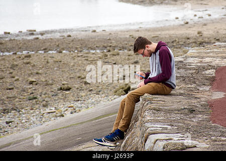 Mann sitzt auf Leiste mit seinem Handy. Am Meer im Hintergrund. Beaumaris Strand, Anglesey, North Wales, UK., 21. April 2017. Stockfoto