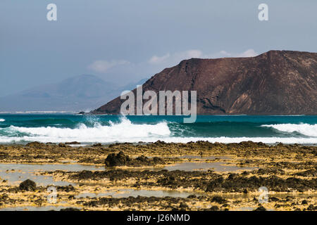 Blick auf Isla de Lobos in der Nähe von Corralejo mit Felsenküste und Surfen im Vordergrund in Fuerteventura, Spanien. Stockfoto