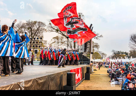 Hinokuni Yosakoi Dance Festival. Team von jugendlichen Frauen tanzen auf der Bühne und trug gemusterten happi Mäntel. Massive rote Flagge geschwenkt werden über asudience. Stockfoto