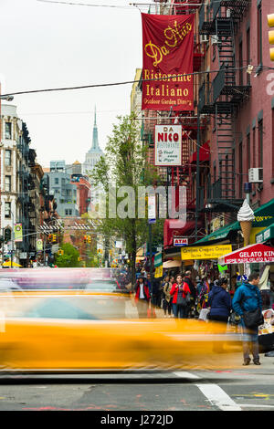 Ein gelbes Taxi fährt vorbei an einer belebten Straße mit Restaurants und Wohnungen mit dem Empire State Building im Hintergrund, Little Italy, New York Stockfoto