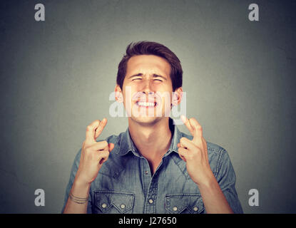 Junger Mann wünschen seine Daumen drücken isoliert auf graue Wand Hintergrund. Besonderer Moment warten Stockfoto