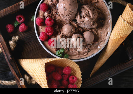 Schüssel mit hausgemachten Schokoladeneis mit frischen Himbeeren, Minze und Waffel Kegel auf Holz Tablett über schwarzen verbrannten Holz Hintergrund. Ansicht von oben, Nahaufnahme Stockfoto