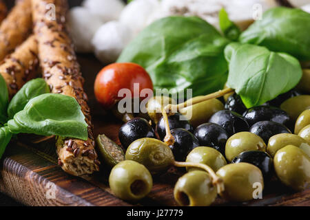 Mediterrane Vorspeise Antipasti-Board mit grünen und schwarzen Oliven, Feta-Käse, Mozzarella, Kapern, Pfeffer, Basilikum mit Grissini Brot über schwarz klebt Stockfoto