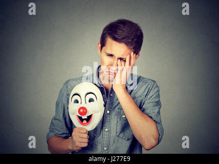 Trauriger Jüngling mit happy Clownsmaske auf graue Wand Hintergrund isoliert. Menschliche Gefühle Stockfoto