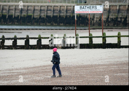 Eine Frau trägt eine warme Jacke, eine Mütze und Handschuhe zu Fuß am Strand entlang an einem kalten Tag im Littlehampton, West Sussex, England. Stockfoto