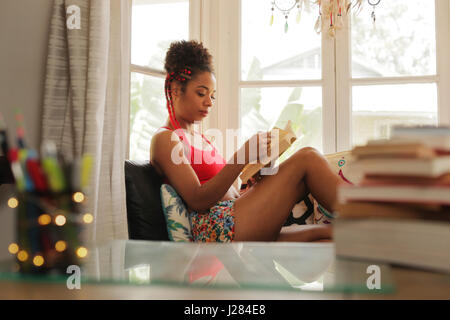 Glückliche Studentin Buch zu lesen und auf Sofa sitzen. Junge afrikanische amerikanische Frau, entspannend, schwarze Mädchen auf der Couch liegen. Hispanische People- und lifestyle Stockfoto