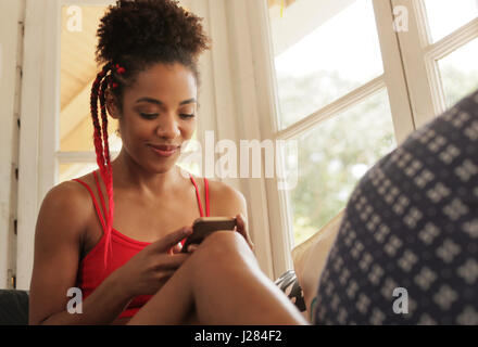 Schwarze Mädchen auf der Couch liegend und mit Smartphone, junge afroamerikanische Frau entspannend mit Handy. Glücklich Latina auf Sofa sitzen, Lächeln und tex Stockfoto