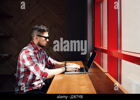 Junger Arbeiter in einem Büro oder Café am Computer sitzen. Freier Mitarbeiter in einem karierten Hemd. Der Designer sitzt vor einem Fenster am Arbeitsplatz. Stockfoto