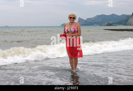 In voller Länge Portrait von ältere Frau, die am Kiesstrand mit Strand-Packung in die Hüften auf Surf-Wellen und Klippe Hintergrund in trübe stürmischen steht Stockfoto