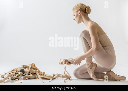 Blonde Ballett-Tänzerin mit geschlossenen Augen auf dem Boden sitzt und hält einen Pointe Schuh auf dem hellen Hintergrund im Studio. Sie trägt einen Beige Tanz-Verschleiß. N Stockfoto