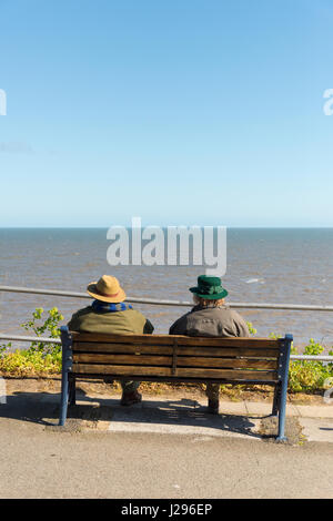 Ansicht von hinten von zwei Personen in Hüte und Mäntel bei sonnigem Wetter sitzt auf einer Sitzbank mit Blick auf das Meer bei Cromer Norfolk UK Stockfoto
