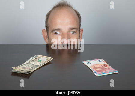 Porträt des kaukasischen erwachsenen Mannes Gesicht blickte über dunklen braunen Tisch. Ein Auge, die in Dollar und andere an Euro-Banknoten Haufen wie Squint Augen oder Stockfoto