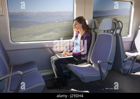 braune Haare lächelnde Frau gekleidet in lila und blau, Sitzung, die Anreise mit der Bahn, die Eingabe von Stichwort Laptop-Computer, stützte sich auf ihre Beine Stockfoto