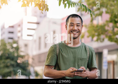 Junger asiatischer Mann, der in die Kamera lächelt, während er eine SMS auf seinem Handy schickt. Draußen, städtischer Hintergrund, eingerahmt von Ästen und Blättern. Stockfoto