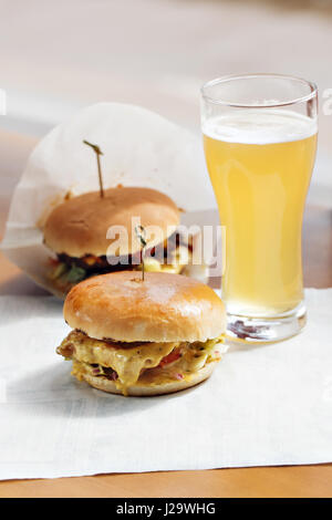 Zwei leckere Burger, garniert mit Zwiebeln, Tomaten, Cheddar-Käse-Scheibe und Kopfsalat mit Nacho-Chips auf Schneidebrett und Glas Bier im Hintergrund gut gemacht Stockfoto