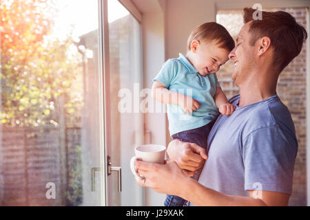Vater hält kleinen Sohn beim Kaffeetrinken zu Hause am Fenster Stockfoto