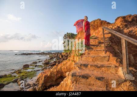 Junge Frau, gekleidet in einem roten Seide Sari steht an einem tropischen Paradiesstrand in Mirissa, Sri Lanka. Stockfoto