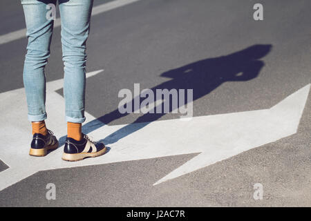 Seitenansicht Nahaufnahme von einer jungen Frau mit schwarzen Schuhen und blaue Jeans stehen auf einer Straße mit Pfeil-Zeichen zeigen in verschiedene Richtungen Konzept Stockfoto