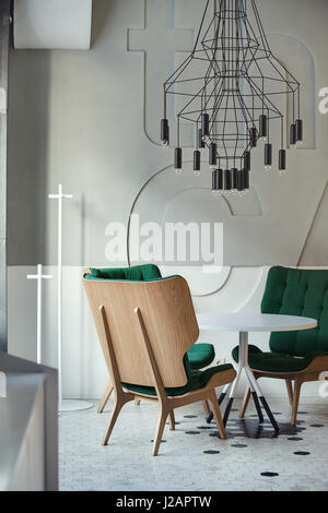Drei grüne Sessel um weißen runden Tisch im Café in einem modernen Stil mit hellen Wänden. Über den Tisch und schwarz hängen schwarze Lampen Stockfoto